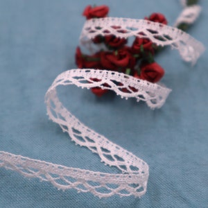 Może przedstawiać: Biała koronka z delikatnym falbaniastym brzegiem. Koronka jest pokazana na niebieskim tle z czerwonymi jedwabnymi kwiatami.