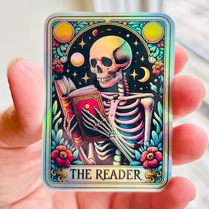 May include: A holographic tarot card with a skeleton reading a book. The card is decorated with flowers and stars. The text on the card reads "The Reader".