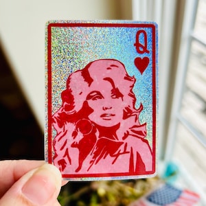 May include: A holographic sticker featuring a red illustration of a woman's face, styled as a playing card. The card is the Queen of Hearts, with a red heart symbol and the letter "Q" in the upper right corner. The background has a shimmering, iridescent effect.
