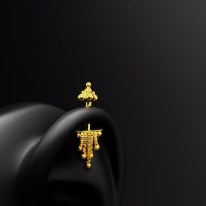 Op de afbeelding: Een paar gouden oorbellen met een uniek ontwerp. Eén oorbel is een kleine knop met een cluster van ronde accenten. De andere heeft een hangend ontwerp met een reeks kleine, ronde accenten.