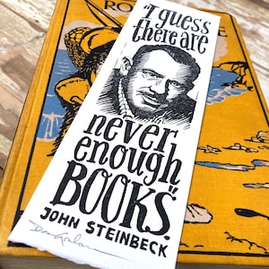 Op de afbeelding: Een witte bladwijzer met zwarte tekst en een zwart-witte illustratie van John Steinbeck. De tekst luidt "Ik denk dat er nooit genoeg BOEKEN zijn" en is ondertekend met "John Steinbeck".