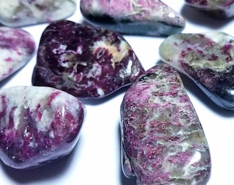 May include: A collection of polished gemstones with a mix of colors, including deep pink, white, and gray. The stones have a smooth, rounded shape and a natural, organic appearance. The gemstones are scattered across a white surface.