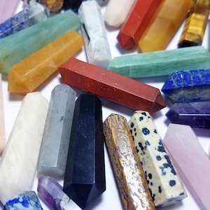 May include: Assortment of colorful gemstone wands in various colors, including red, green, yellow, blue, and black. The wands are hexagonal and vary in size. The gemstones are arranged on a white surface.