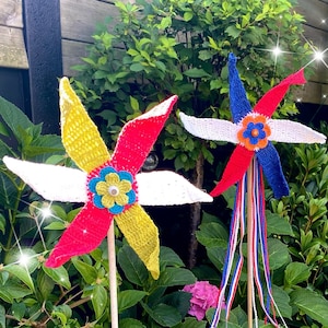 Op de afbeelding: Twee kleurrijke gehaakte windmolens met witte, rode, blauwe en gele accenten. De windmolens zijn bevestigd aan houten stokken en staan in een tuin met groen gebladerte en roze bloemen.