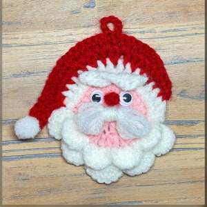 Op de afbeelding: Een gehaakte kerstversiering van een kerstman met een rode muts, witte baard, roze wangen en zwarte ogen.