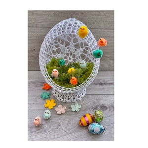 Può includere: Un cestino a forma di uovo in crochet bianco con una base di muschio verde e pulcini colorati all'interno. Ci sono anche quattro fiori all'uncinetto e tre uova di cioccolato avvolte su una superficie di legno grigia.