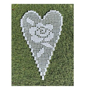 Op de afbeelding: Wit gehäkeld hartvormig doily met een delicaat bloemenpatroon, getoond op een groene grasachtige ondergrond.