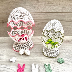 Op de afbeelding: Twee gehaakte paaseierenmandjes, een wit met roze rand en de andere wit met groene rand. Beide mandjes zijn gevuld met kleine chocolade-eieren. Er zijn ook drie gehaakte konijnenvormen in roze, wit en groen.