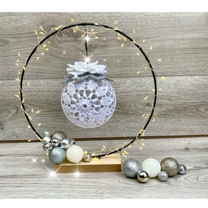 May include: A white crocheted Christmas ornament with a silver flower detail hangs from a black metal hoop decorated with a string of lights. The hoop is on a wooden base with silver and gold ornaments.