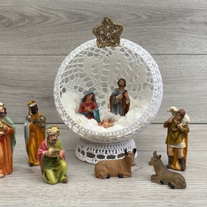 Op de afbeelding: Een wit gehaakte kerststal met een ster bovenop. De scène bevat Maria, Jozef, baby Jezus, drie wijzen, een herder, een schaap, een koe en een ezel.