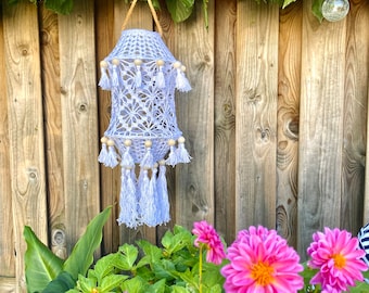 May include: A white crocheted lantern with wooden beads and tassels hanging from a wooden fence. The lantern is surrounded by pink flowers and green leaves.