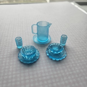 May include: A miniature blue glass tea set, including a pitcher on a saucer, two small glasses, and two plates. The set is made of translucent blue glass and has a delicate, vintage appearance. The pieces are arranged on a light gray surface.