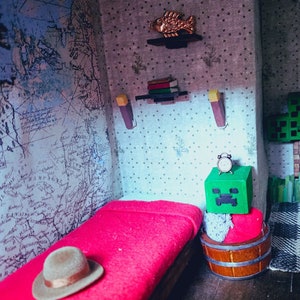 May include: A miniature bedroom with a red bed, a green Minecraft Creeper block with a clock on top, a wooden barrel, and a brown hat on the bed.