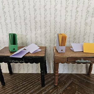 May include: Miniature desks with file organizers and papers. One desk has a black base and the other has a brown base. The file organizers are green and purple, and the papers are yellow and purple. The desks are made of wood.