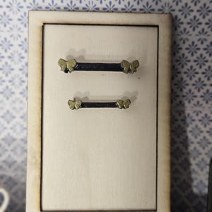 May include: A wooden cabinet door with two black handles, each adorned with gold butterfly accents. The cabinet door is set against a light background, with a decorative blue and white floral wallpaper in the background.