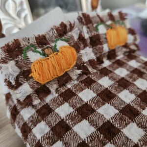 May include: Miniature bed set with a brown and white checkered pattern. The bedding features embroidered orange pumpkins with green stems. The bed set includes a pillow and a blanket, perfect for a dollhouse or seasonal decor.