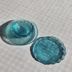 May include: Two clear, light blue glass objects with scalloped edges. One is a small, domed shape set on a larger, flat base. The other is a flat, round dish. The objects are on a pink and white patterned surface.