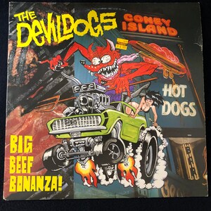 May include: Album cover for "The Devil Dogs" with a cartoon devil driving a green hot rod. The cover features the text "Coney Island Hot Dogs" and "Big Beef Bonanza!" in a bold, yellow font.