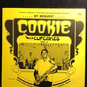 Pode incluir: Uma capa de álbum de vinil vintage com fundo amarelo e texto preto. O álbum é intitulado "COOKIE & THE CUPCAKES" e apresenta uma foto de um homem tocando saxofone. O texto diz "Another Collector's Item From The Louisiana Bayou Country".