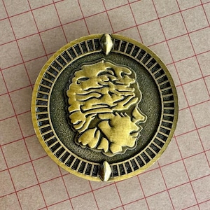 May include: A round, antique gold-colored coin with a detailed profile of a person and a brain-like design. The coin has a textured border and is set against a grid background.