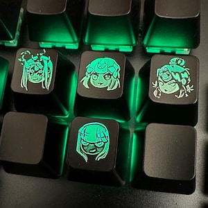 May include: Black keyboard keys with green backlighting. Four keys feature glowing green anime-style character designs. The characters have different hairstyles and facial expressions. The keyboard is a gaming keyboard.