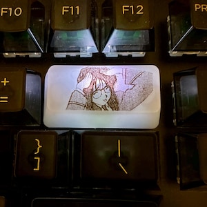 Puede incluir: Una tecla de teclado mecánico blanca translúcida que presenta una ilustración de línea negra de un personaje de estilo anime con gafas, con las manos sobre la cabeza, colocada entre teclas negras etiquetadas con F10, F11, F12 y ENTER.
