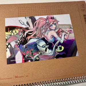 Op de afbeelding: Een kleurrijke anime-stijl print met een personage met roze haar, een bril en een zwarte hoed. De print bevat een beerachtig personage met een bril en de tekst "DANGANRONPA". De print staat op een spiraalgebonden notitieboek.