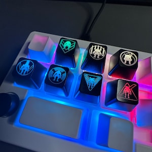 Könnte beinhalten: Ein Satz von acht Tastenkappen mit verschiedenen Designs, darunter ein Roboter, ein Schädel und ein geometrisches Muster. Die Tastenkappen sind mit blauem und grünem Licht beleuchtet.