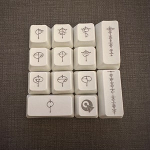 May include: A set of 10 white keycaps with various symbols and designs. The keycaps are arranged in a 3x3 grid with a single keycap below the grid. The keycaps are all white with black symbols and designs. The symbols and designs include a cross, a circle with a line through it, a circle with a dot in the centre, and a circle with a line through it and a dot in the centre.