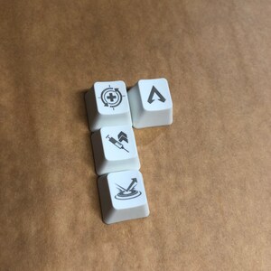 May include: Four white keycaps with different symbols printed on them. The symbols are a crosshair with a plus sign, a syringe with an arrow pointing up, an arrow pointing up with a circle around it, and a triangle with an arrow pointing up.