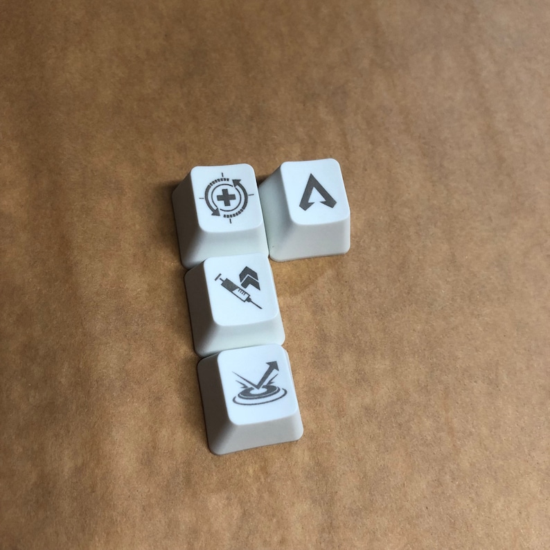 May include: Four white keycaps with different symbols printed on them. The symbols are a crosshair with a plus sign, a syringe with an arrow pointing up, an arrow pointing up with a circle around it, and a triangle with an arrow pointing up.