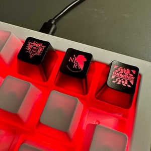 May include: Three black keyboard keycaps with different designs. The first keycap has a red and white logo with the text "NERV". The second keycap has a red and white logo with the text "NERV". The third keycap has a red and white logo with the text "EMERGENCY" in English and Japanese.
