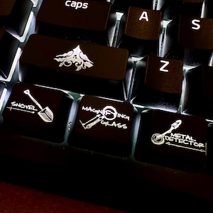 May include: A close-up of a black keyboard with illuminated keys. The keys display white illustrations of a shovel, magnifying glass, and metal detector. Other keys include the letters "A", "S", and "Z", and the word "caps".