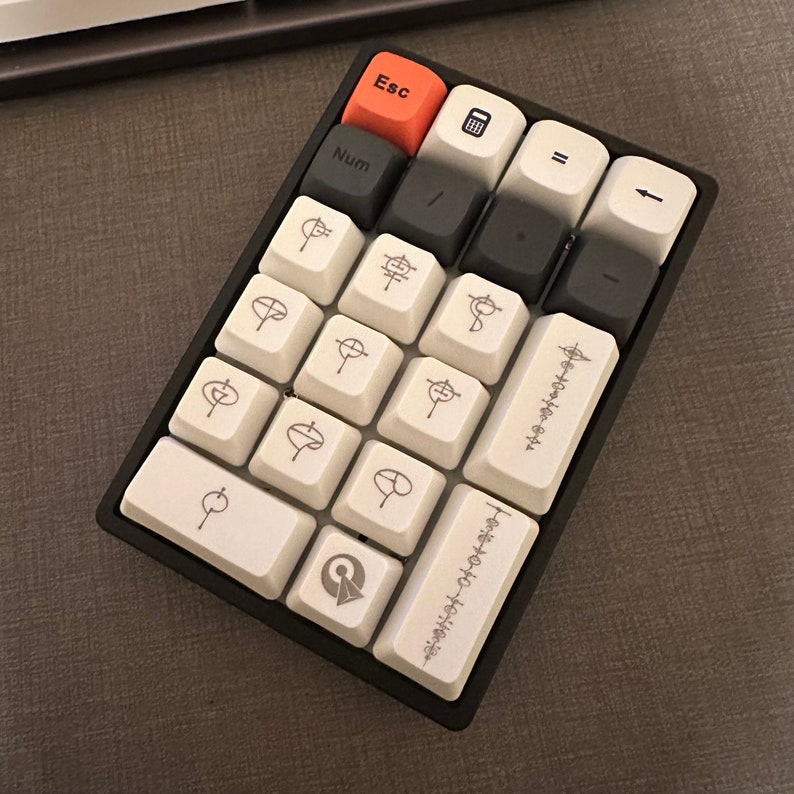 May include: A black and white mechanical keyboard with a red escape key. The keyboard has a numeric keypad with 12 keys and a few function keys. The keys have a circular design with a symbol on each key.