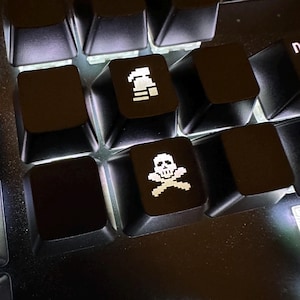 Puede incluir: Primer plano de un teclado mecánico negro con teclas iluminadas. Dos teclas presentan gráficos blancos pixelados: una calavera y huesos cruzados, y una campana. Las otras teclas están en blanco. El teclado está iluminado desde abajo.