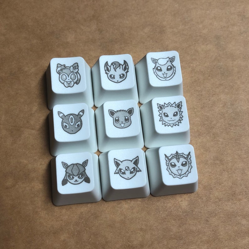 May include: Nine white keycaps with black and gray Pokemon character designs. The characters include Espeon, Umbreon, Flareon, Jolteon, Vaporeon, Leafeon, Glaceon, Sylveon, and  Eevee.
