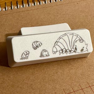 May include: A white rectangular keyboard key featuring a black line drawing of a woodlouse. The drawing includes three smaller woodlice and one larger woodlouse. The key is on a brown surface.
