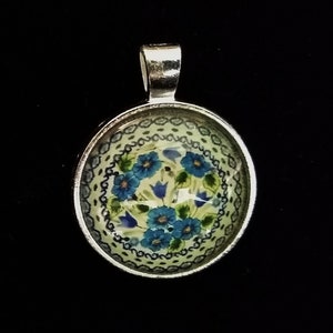 May include: A silver pendant with a round, white and blue floral design. The pendant has a silver bail for attaching to a chain.