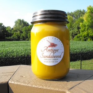 May include: A yellow glass jar candle with a white label that reads "Antique Sandalwood 100% Soy Candle Made in the USA".