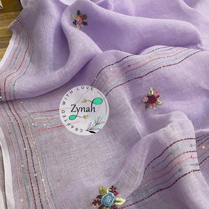 May include: A lavender linen scarf with embroidered flowers and a border of sequins. The scarf is handmade and has the text "Crafted with Love Zynah" in a circle.