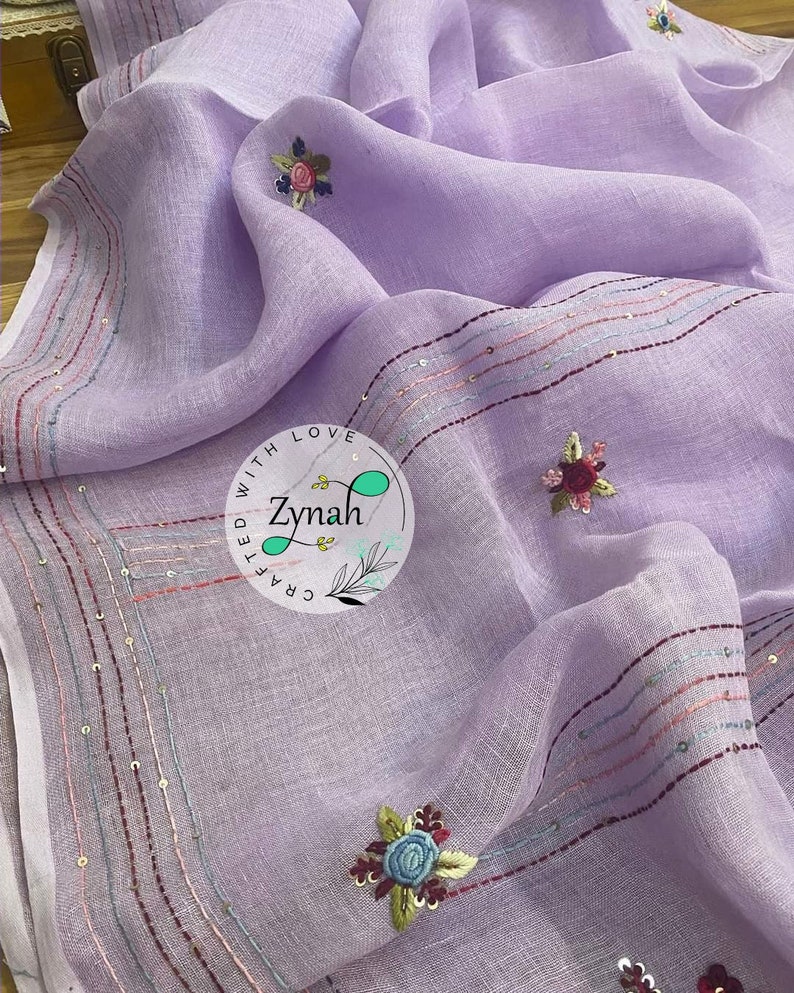 May include: A lavender linen scarf with embroidered flowers and a border of sequins. The scarf is handmade and has the text "Crafted with Love Zynah" in a circle.