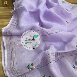 May include: A lavender linen saree with embroidered floral designs and sequins along the border. The saree is crafted with love by Zymah.