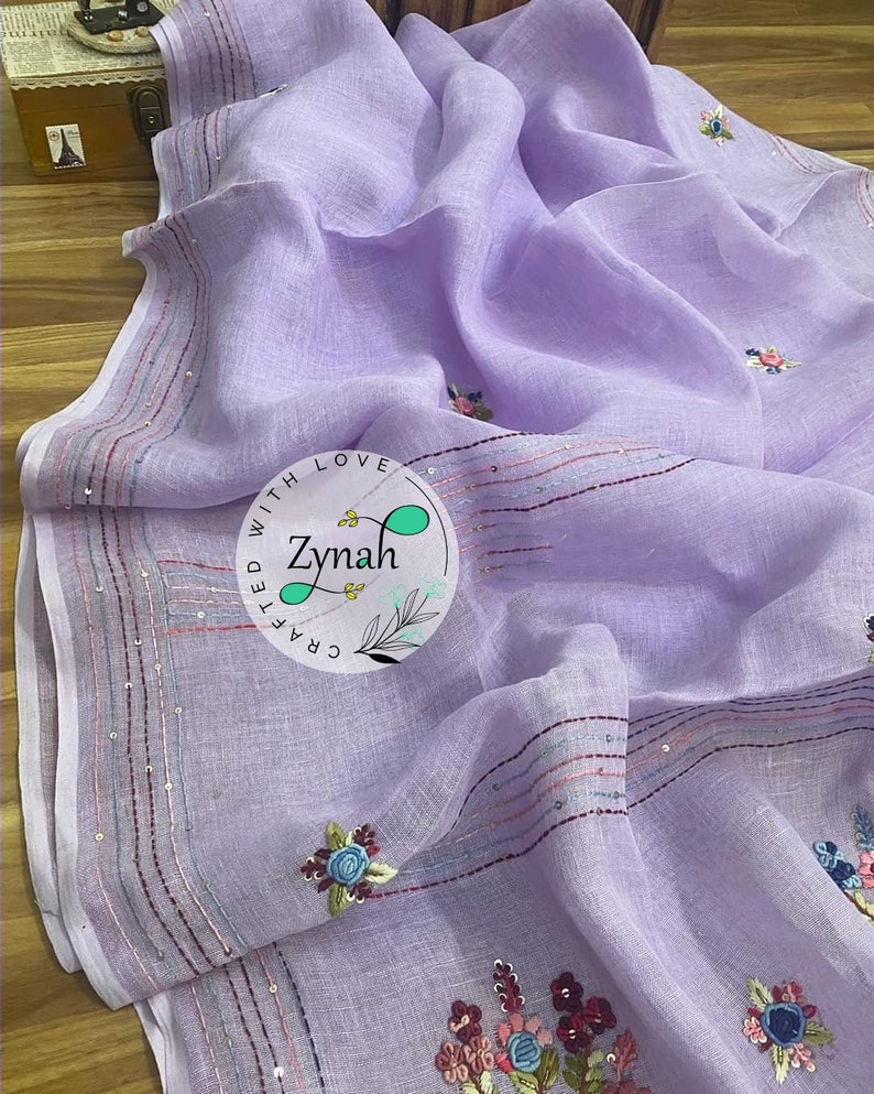 May include: A lavender linen saree with embroidered floral designs and sequins along the border. The saree is crafted with love by Zymah.