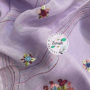 May include: A lavender fabric with embroidered flowers in shades of pink, red, blue, and green. The fabric is decorated with lines of stitching and sequins. The text "Crafted with Love Zynah" is embroidered in a circle on the fabric.