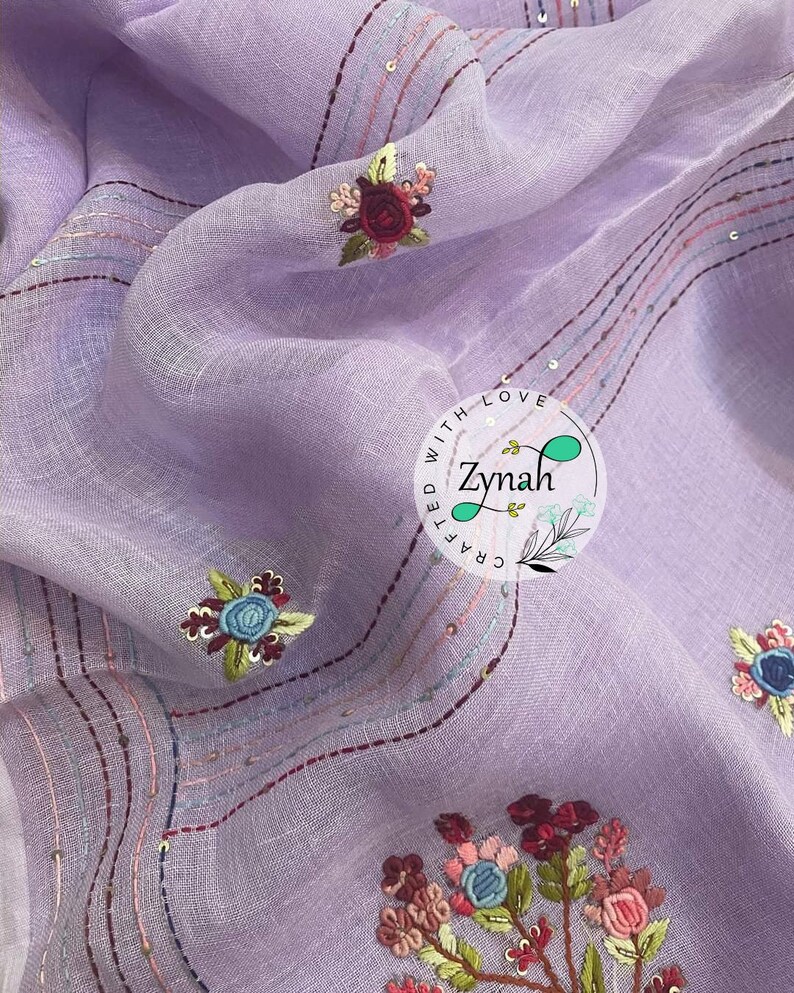May include: A lavender fabric with embroidered flowers in shades of pink, red, blue, and green. The fabric is decorated with lines of stitching and sequins. The text "Crafted with Love Zynah" is embroidered in a circle on the fabric.