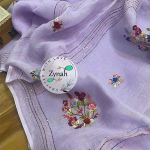 May include: A lavender linen scarf with embroidered floral designs and sequins. The scarf has a border of pink, blue, and white stripes. The text "Crafted with Love Zynah" is embroidered on the scarf.