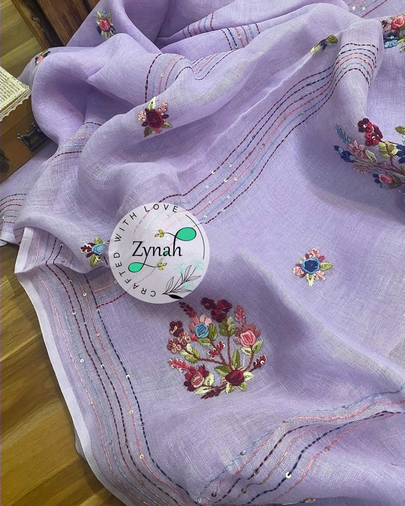 May include: A lavender linen scarf with embroidered floral designs and sequins. The scarf has a border of pink, blue, and white stripes. The text "Crafted with Love Zynah" is embroidered on the scarf.