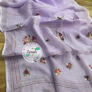 May include: A lavender linen saree with embroidered floral designs and a border of sequins and stripes. The saree has a label that says "Crafted with Love Zynah".