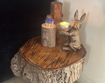 May include: A live edge wood slice side table with black metal hairpin legs. On the table sits a wooden vase with a red flower, a wooden matchstick holder with blue-tipped matches, and a bronze-colored rabbit figurine holding a tea light candle.