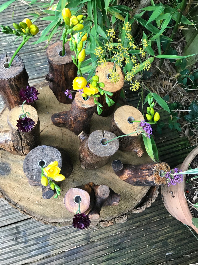 May include: A collection of rustic wooden log vases with holes drilled in the top, arranged on a wooden slice. The vases are filled with yellow and purple flowers.